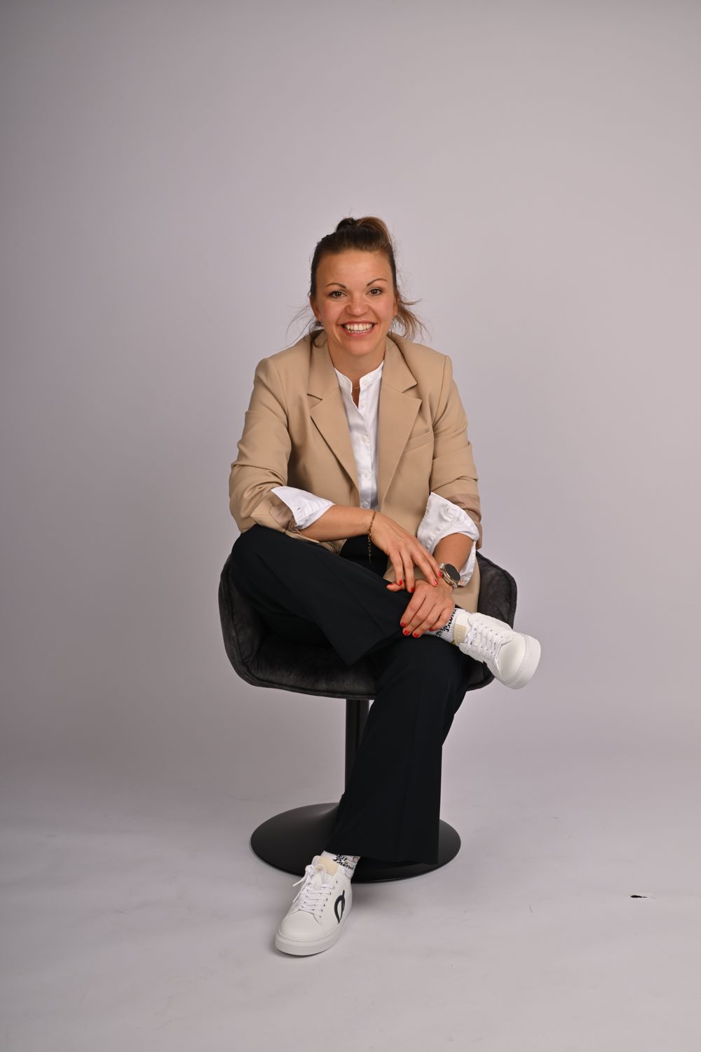 Steffi Reith im beigen Blazer, lächelnd mit überschlagenen Beinen auf einem Stuhl sitzend